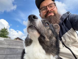 A black and white corgi cardigan and a beared man (me!) photographed from below. The dog is looking up and the man is looking down. There are some clouds and a blue sky in the background.